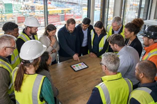 Construction site team gathered around a table reviewing a tablet during a group briefing, with the construction site visible through the windows behind them